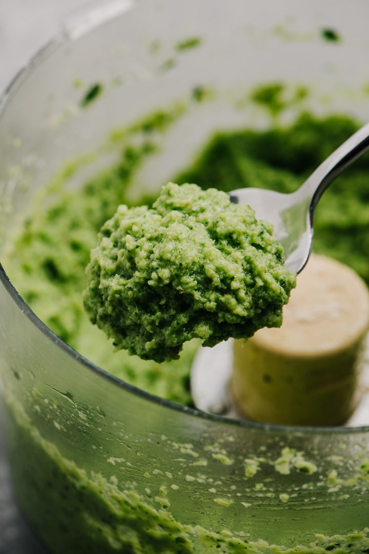A spoonful of broccoli pesto hovering over the bowl of a food processor.