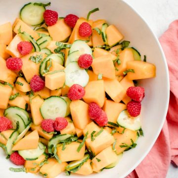 A large serving bowl of cantaloupe melon salad with cucumbers, raspberries, fresh herbs, and a lemon honey dressing.