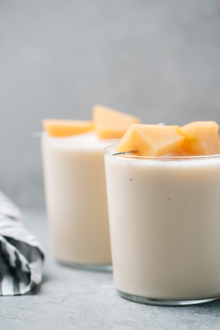 Two cantaloupe and banana smoothies on a cement background topped with fresh cantaloupe. 