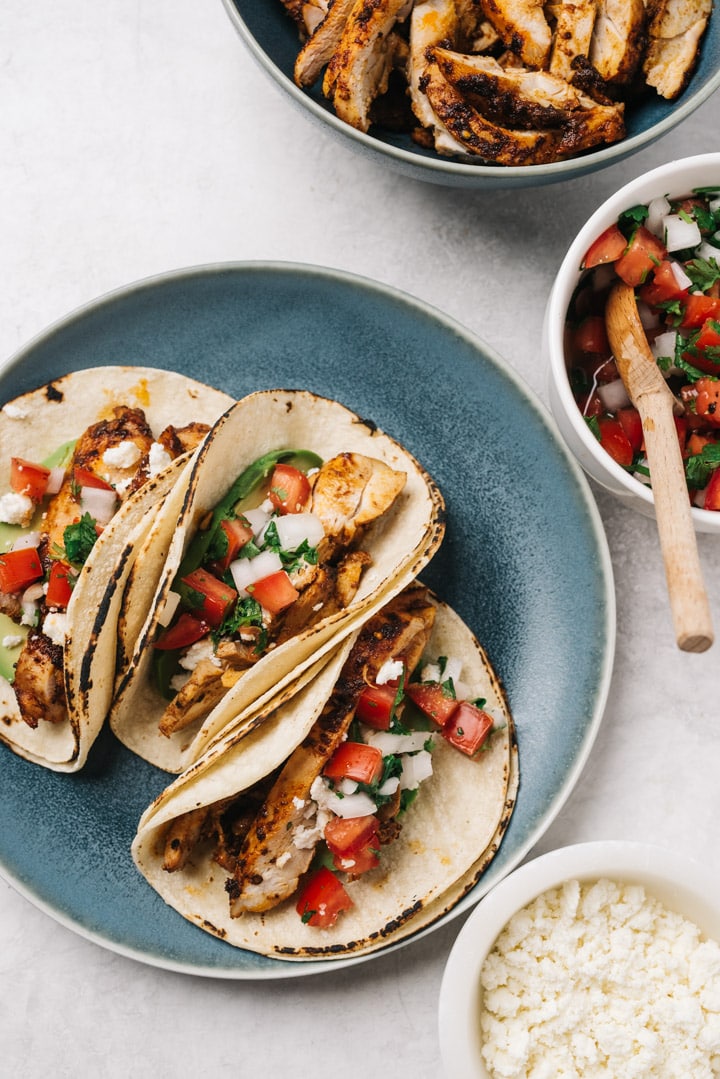 Three chicken street style tacos on a blue plate surrounded by bowls of garnishes and a bowl of cooked chicken slices.