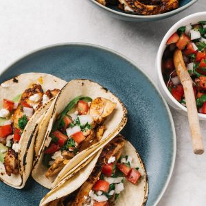 Three chicken street style tacos on a blue plate surrounded by bowls of garnishes and a bowl of cooked chicken slices.