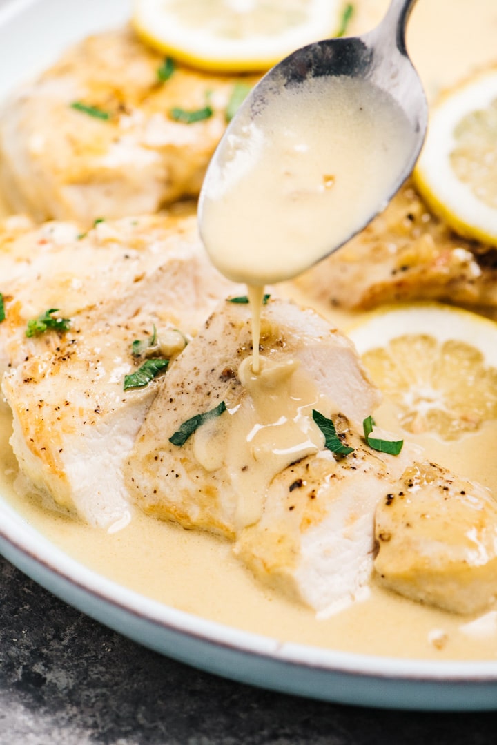A spoon drizzling creamy lemon sauce over a sliced chicken breast on a blue plate.