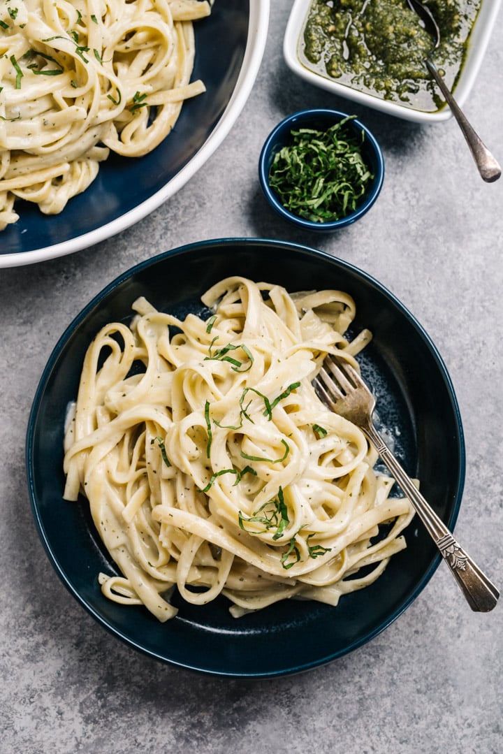 A blue pasta bowl of creamy pesto pasta tossed with fettuccine noodles on a dinner table with small bowls of fresh basil and pesto.