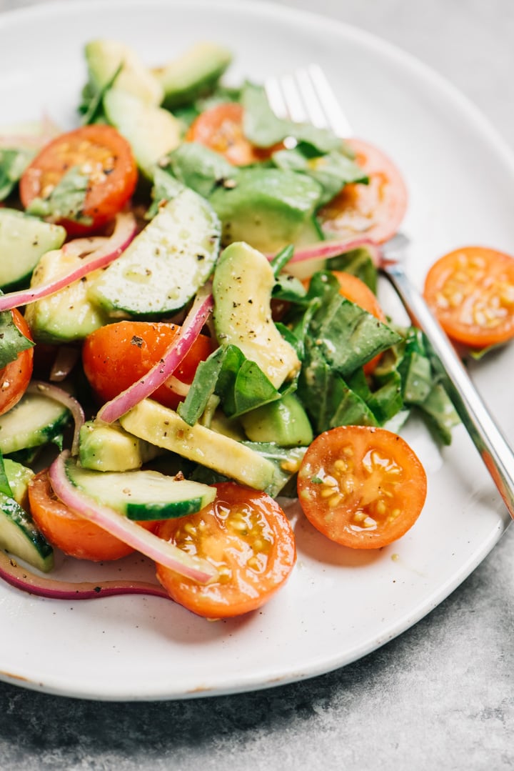 Side view, cucumber and tomato salad with avocado and red onion on a white plate with a fork.
