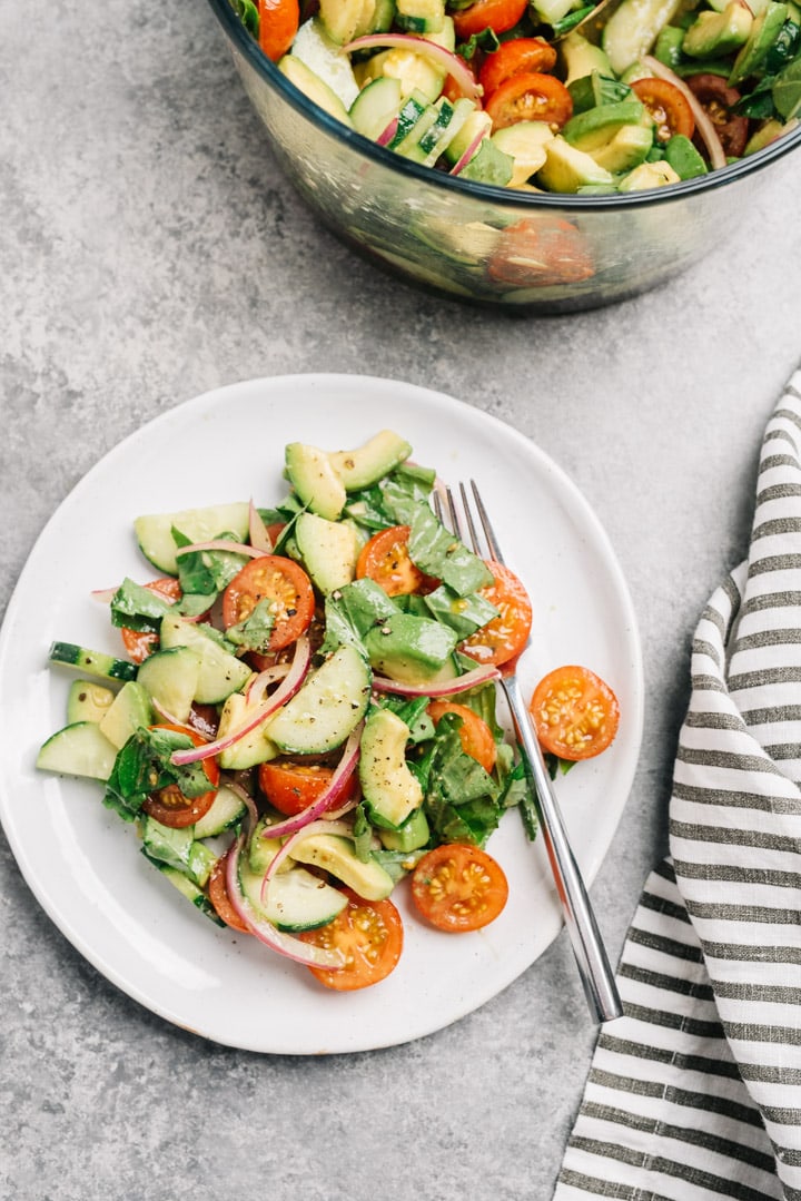 A serving of cucumber and tomato salad with avocado on cement background with a large glass salad bowl and striped linen.