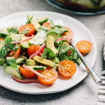 Side view, a serving of cucumber, tomato, and avocado salad on a white plate with a large serving bowl in the background.