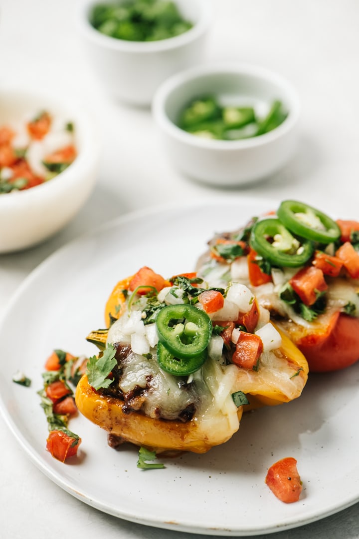 Two keto stuffed peppers on a white plate surrounded with small garnish bowls filled with pico de gallo, cilantro, and sliced jalapenos.