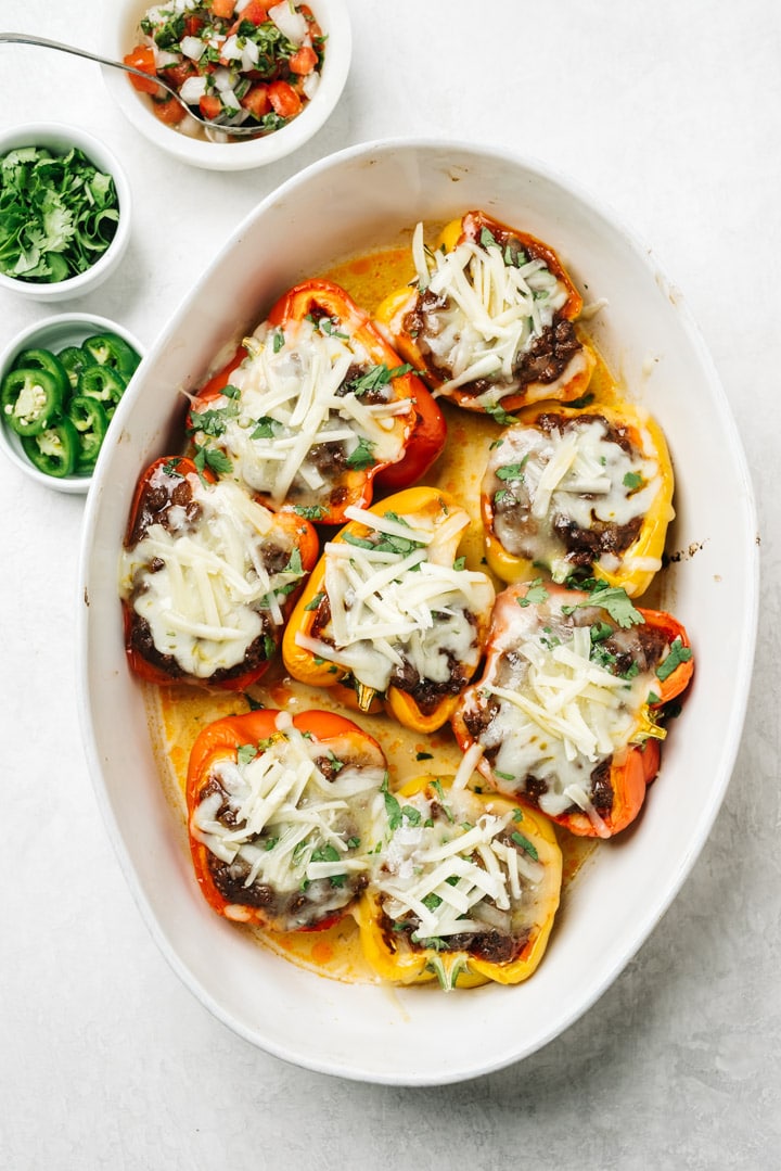 Keto mexican stuffed peppers in a white casserole dish surrounded by small garnish bowls filled with pico de gallo, cilantro, and sliced jalapenos.