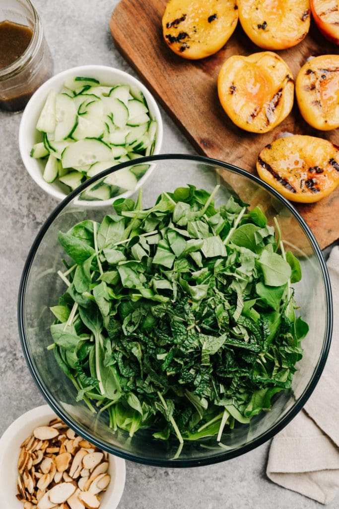 Salad greens with basil and mint in a glass bowl surrounded with grilled peaches, cucumbers, toasted almonds, and balsamic vinegar.
