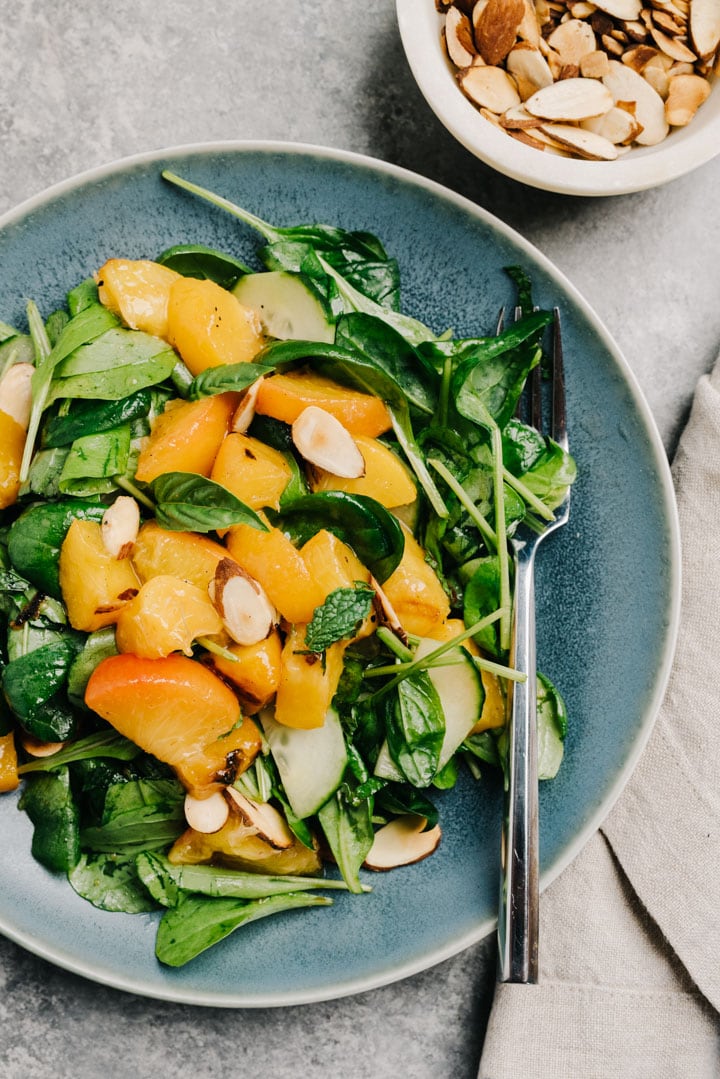 A serving of peach salad with mixed greens, grilled peaches, cucumber, and balsamic dressing on a cement background.