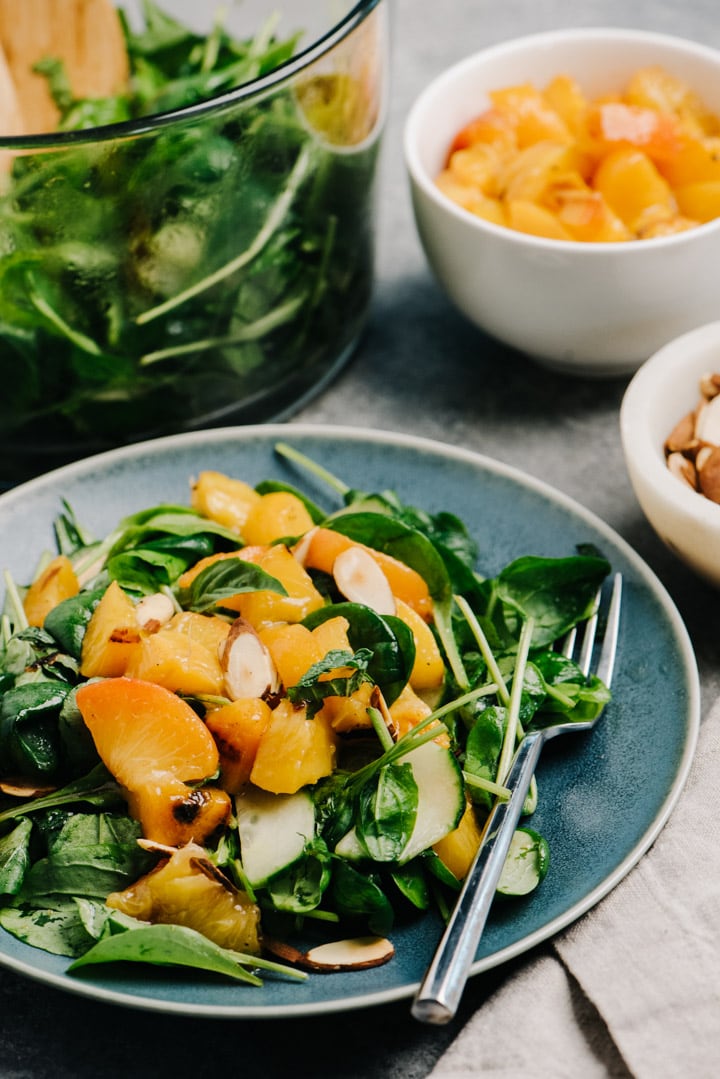 A grilled peach salad on a blue plate with a fork, surrounded by a large bowl of salad greens, diced peaches in a bowl, and a tan linen napkin.
