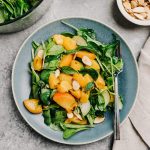 A plate of grilled peach salad on a cement table with a bowl of mixed greens, a bowl of diced grilled peaches, and a small pinch bowl of toasted almonds.