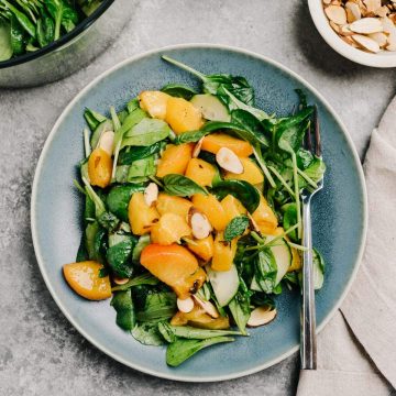 A plate of grilled peach salad on a cement table with a bowl of mixed greens, a bowl of diced grilled peaches, and a small pinch bowl of toasted almonds.