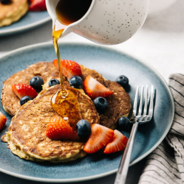 A woman's hand pouring maple syrup onto three banana oatmeal pancakes topped with fresh berries.