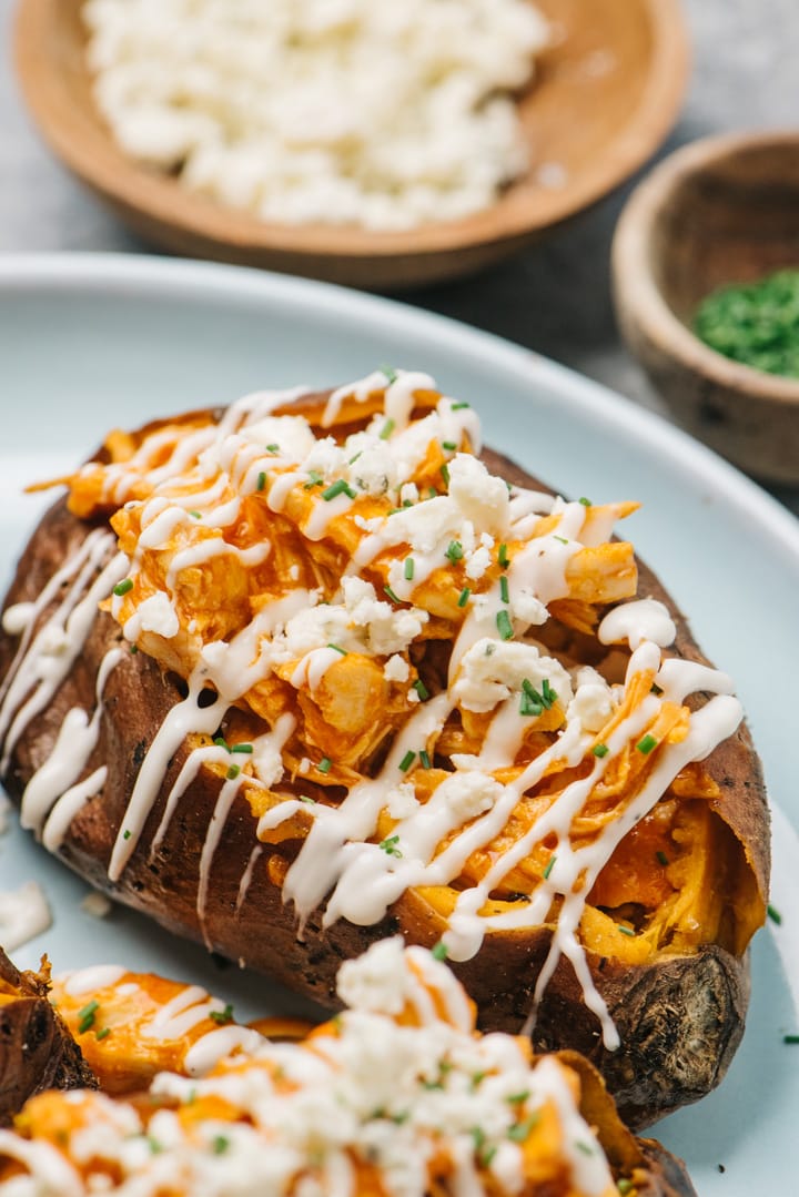 Side view, buffalo chicken stuffed sweet potatoes on a blue plate with small bowls of blue cheese crumbles and chopped chives in the background.