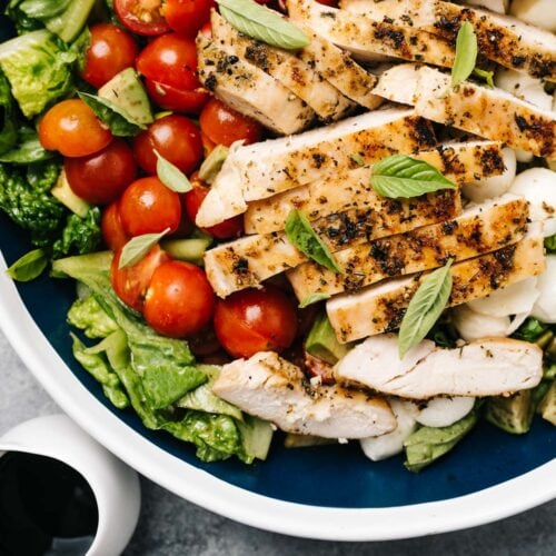 A large serving bowl of caprese salad topped with grilled chicken and a small pitcher of balsamic reduction dressing.