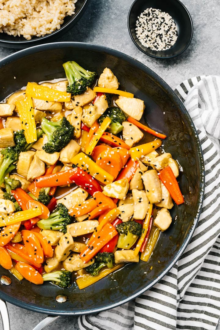 Overhead, healthy chicken stir fry in a skillet with a striped linen napkin, pinch bowl of sesame seeds, and serving bowl of steamed brown rice.