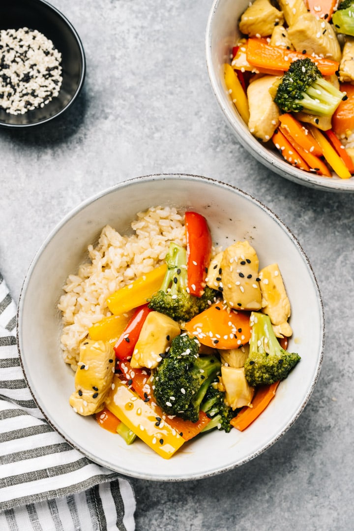 Two bowls of chicken stir fry on a cement background with a small bowl of sesame seeds and a striped linen napkin.