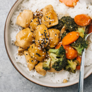 A bowl of chicken teriyaki with white rice and vegetables on a cement background.