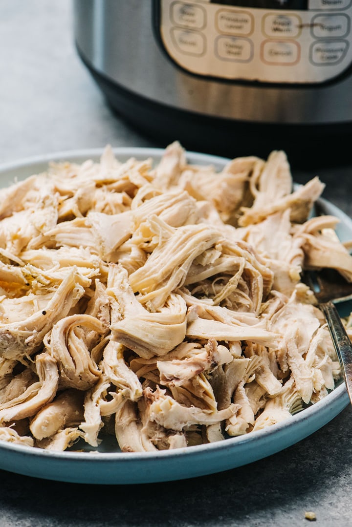 Side view, shredded chicken on a blue platter in front of an instant pot.