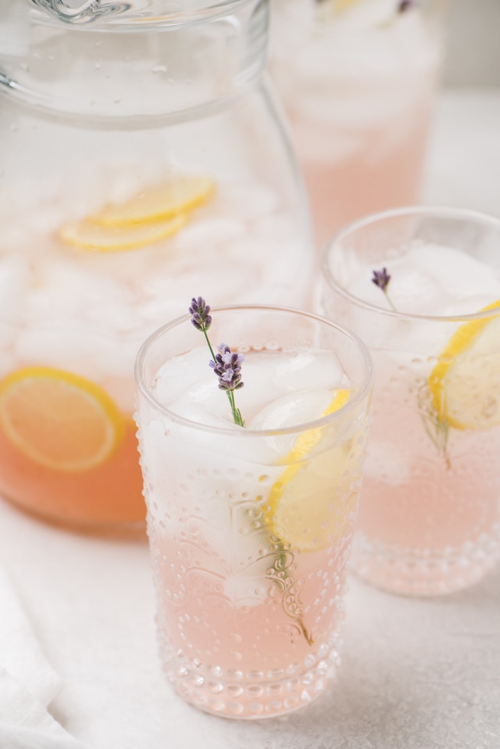A pitcher of lavender lemonade surrounded by several full glasses on a cement background.