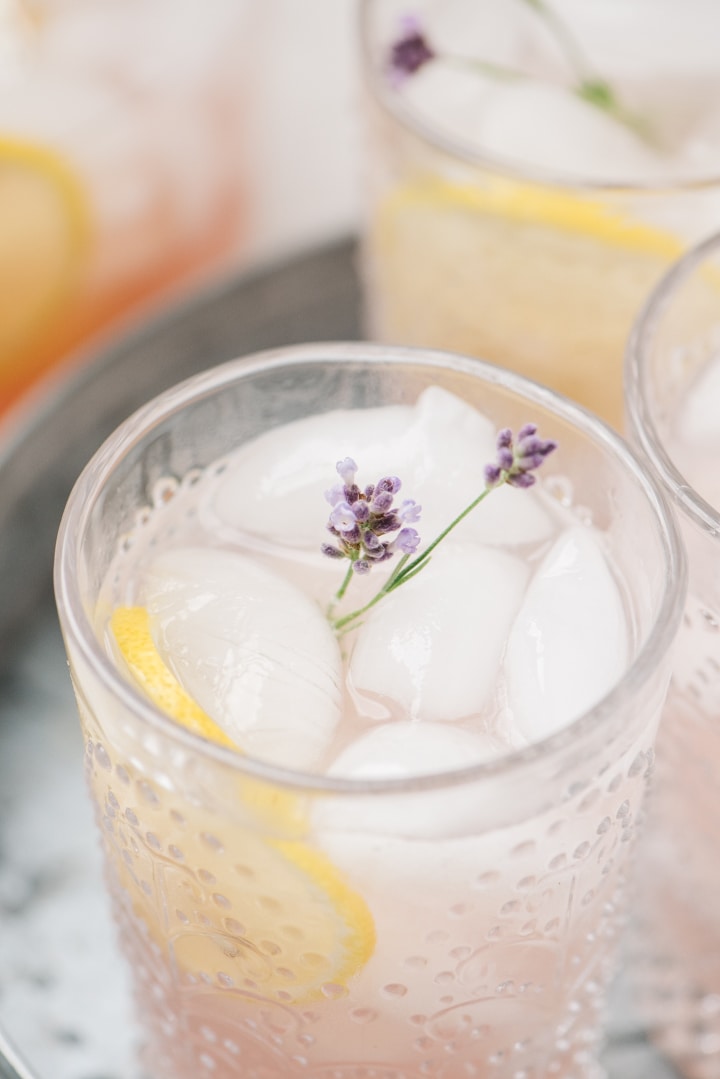 A glass of lemonade made with lavender syrup on a galvanized tray with several glasses in the background.