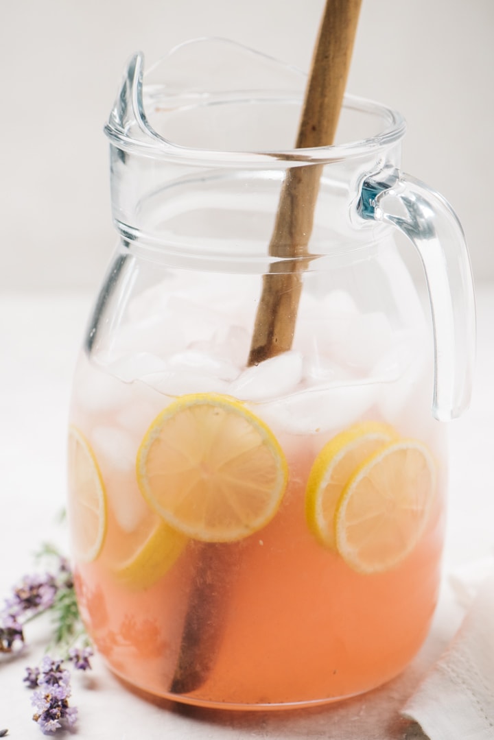 A pitcher of lavender lemonade on a cementer background with lemon slices and fresh lavender flowers.