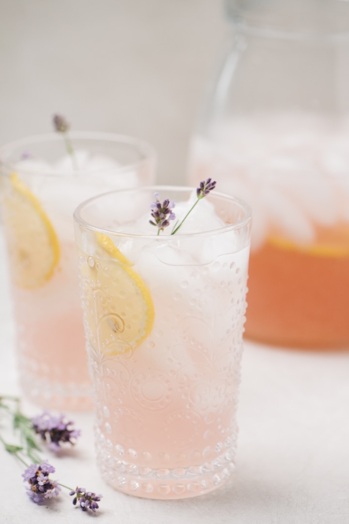 Two glasses of lavender lemonade garnish with lemon slices and lavender flowers, with a pitch of lemonade in the background.