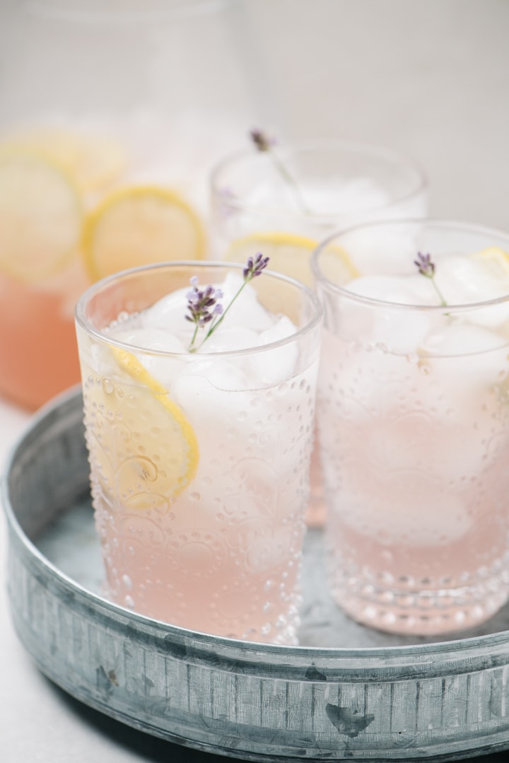 Three glasses of lavender lemonade on a galvanized steel tray, with a pitcher in the background.
