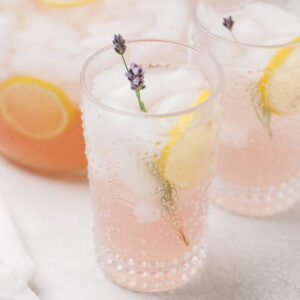 A pitcher of lavender lemonade surrounded by several full glasses on a cement background.