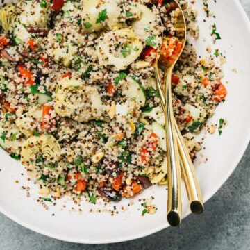 A large mixing bowl of vegan quinoa salad with mediterranean style vegetables including artichoke hearts, olives, and roasted red peppers.