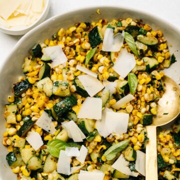 A serving bowl of sauteed zucchini and corn garnished with shaved parmesan and fresh basil.