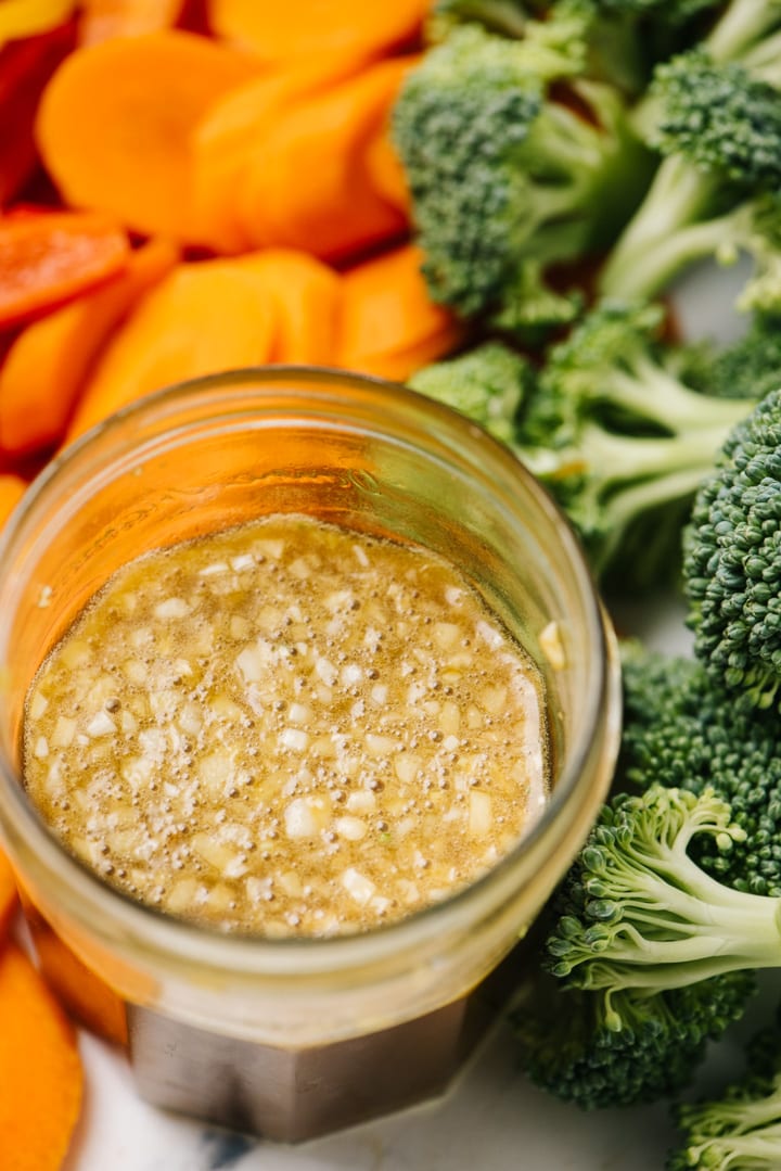 A jar of homemade stir fry sauce surrounded with cut broccoli, carrots, and bell pepeprs.