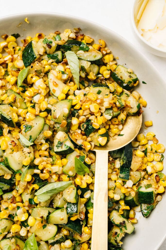 A gold serving spoon resting in a bowl of sauteed zucchini with fresh corn and basil.