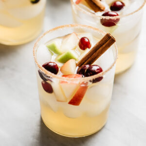 Side view, several glasses of apple cider sangria on a marble table garnished with cinnamon sticks.