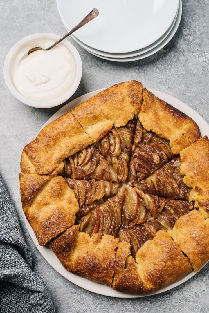 An apple galette on a round white serving platter with a small bowl of vanilla glaze and a dark grey linen napkin.