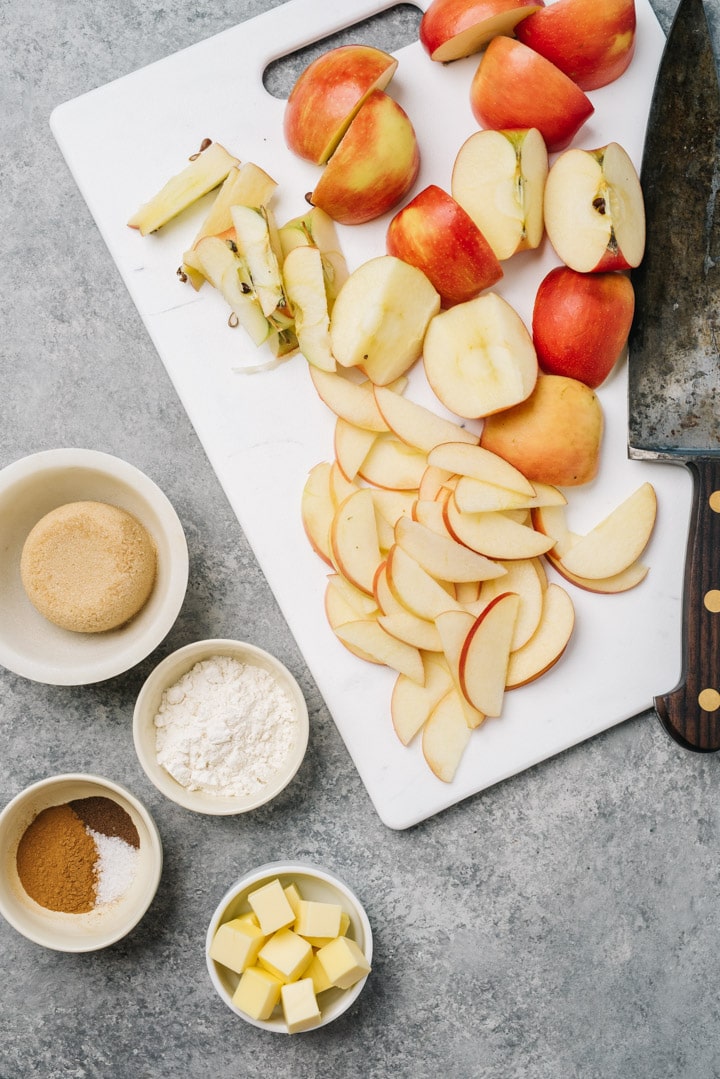 Sliced apples on a white cutting board surrounded with small bowls of brown sugar, spices, flour, and butter.