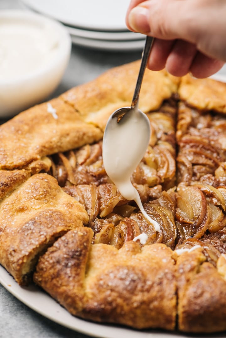 A spoon drizzling vanilla glaze over a sliced apple galette.