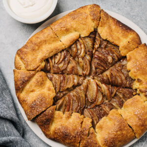 Sliced apple galette on a round serving platter with a bowl of vanilla glaze on the side.