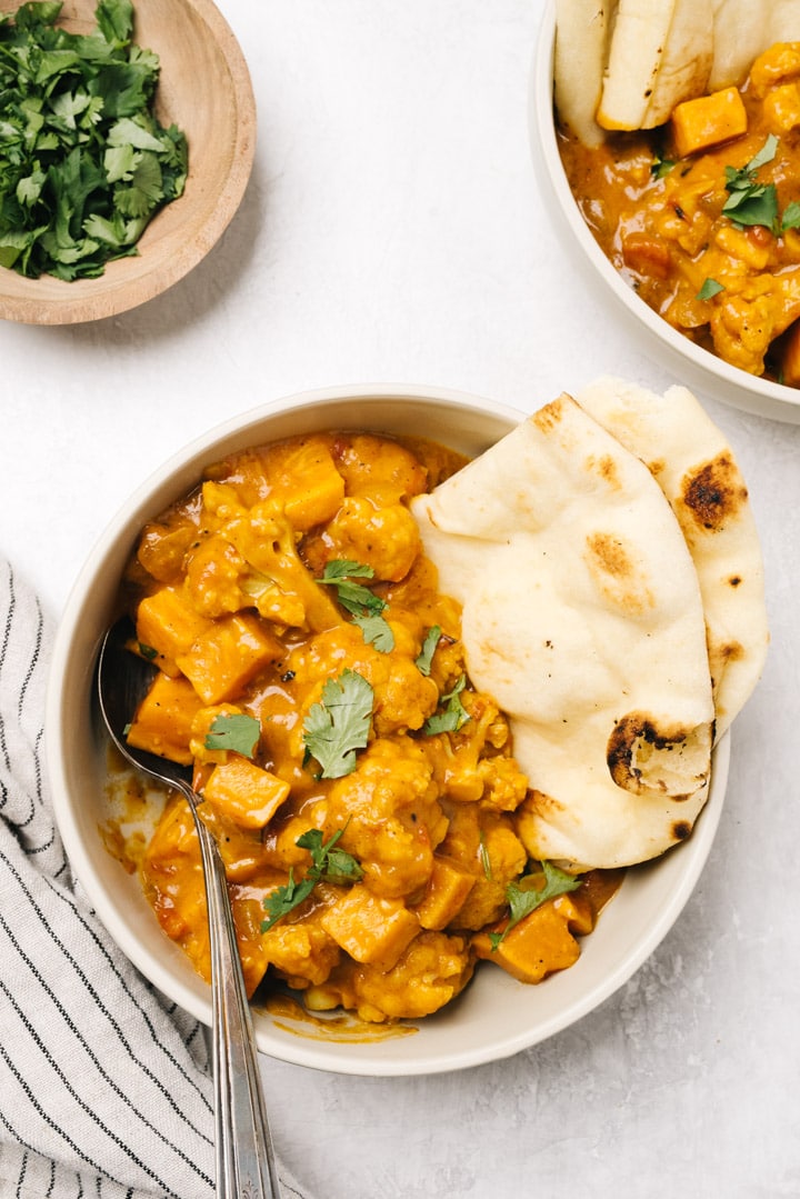 Two bowls of cauliflower curry served with naan bread on a cement background with a small pinch bowl of chopped cilantro.