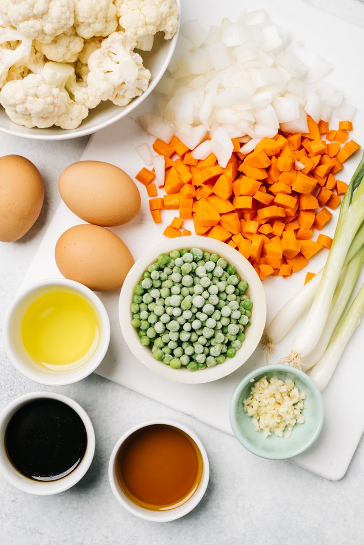 The ingredients for cauliflower fried rice arranged on a cement background - cauliflower, onion, carrot, green onions, peas, ginger, eggs, and seasonings.