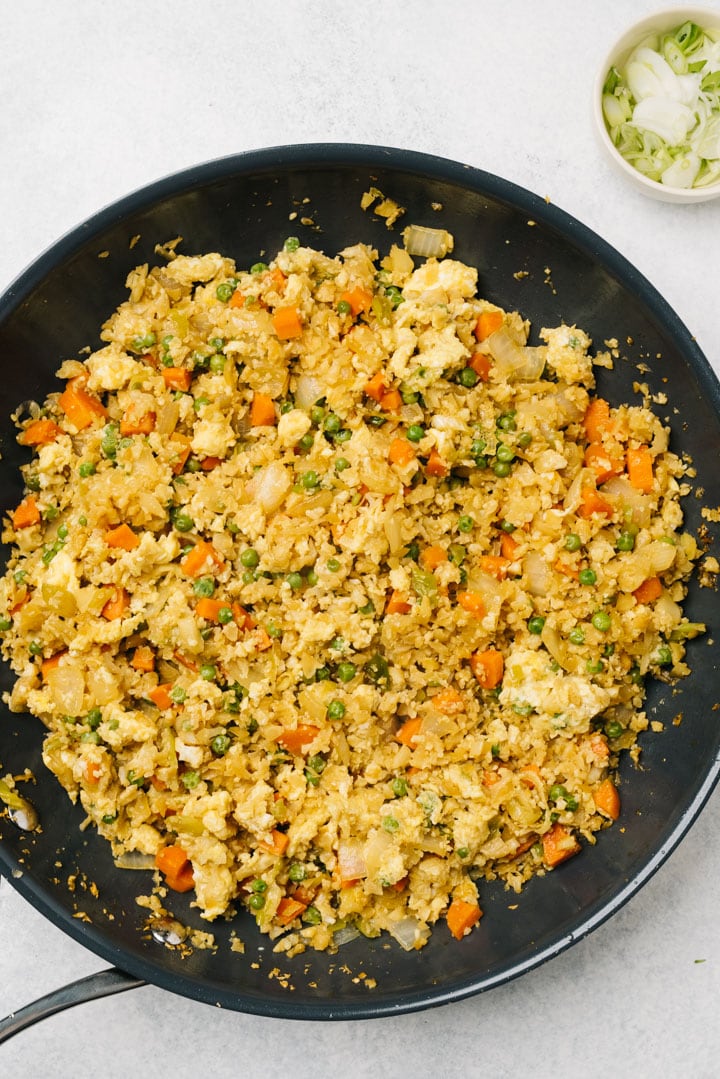 Cauliflower fried rice with in a non-stick skillet with a small bowl of sliced green onions on the side.