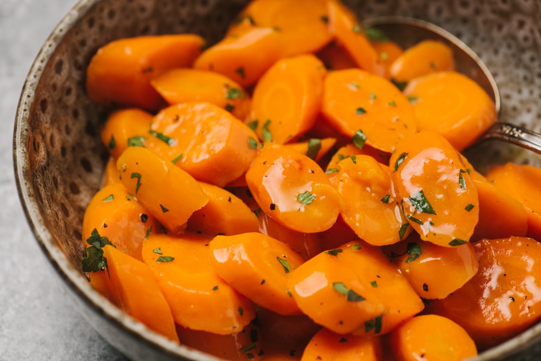 Honey glazed carrots garnished with parsley in a brown serving bowl on a concrete background.
