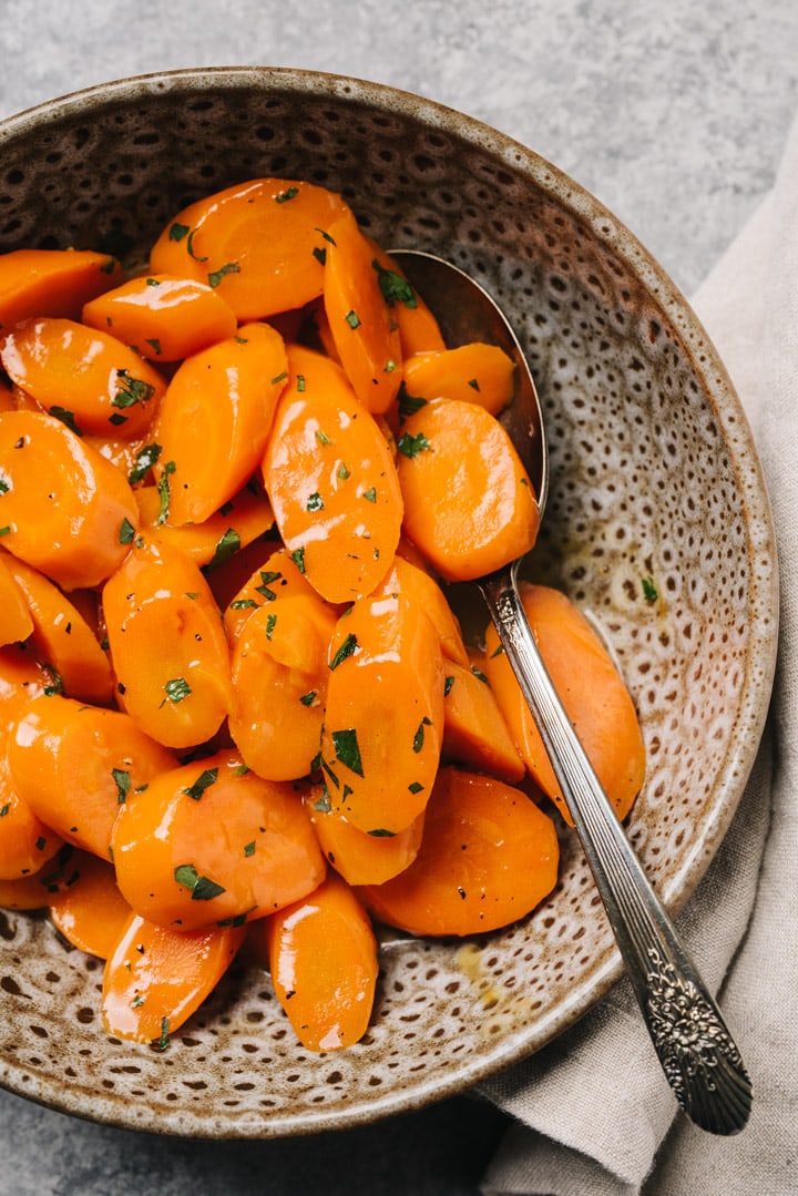 Glazed carrots garnished with parsley in a brown speckled bowl with a vintage serving spoon and tan linen napkin on a concrete background.
