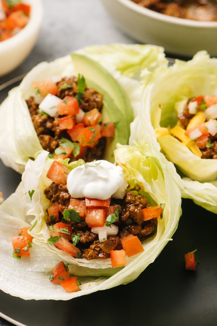 Side view, three ground beef keto tacos in iceberg lettuce shells topped with sour cream, avocado, and pico de gallo.