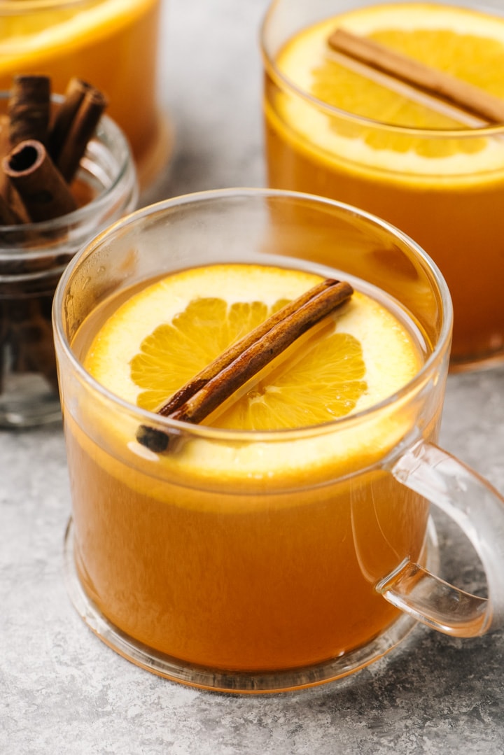 Three glass mugs full of mulled apple cider garnished with orange sliced and cinnamon sticks on a cement table.