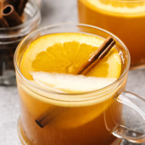 Two mugs of mulled apple cider garnished with an orange slice, apple slice, and cinnamon stick on a cement background.
