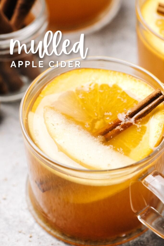 Side view, hot apple cider in a glass mug with an orange slice, apple slices, and cinnamon stick; text overlay reads "mulled apple cider".