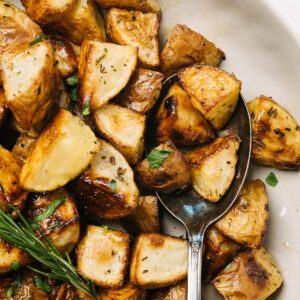 A tan serving bowl of rosemary roasted potatoes with a vintage serving spoon.