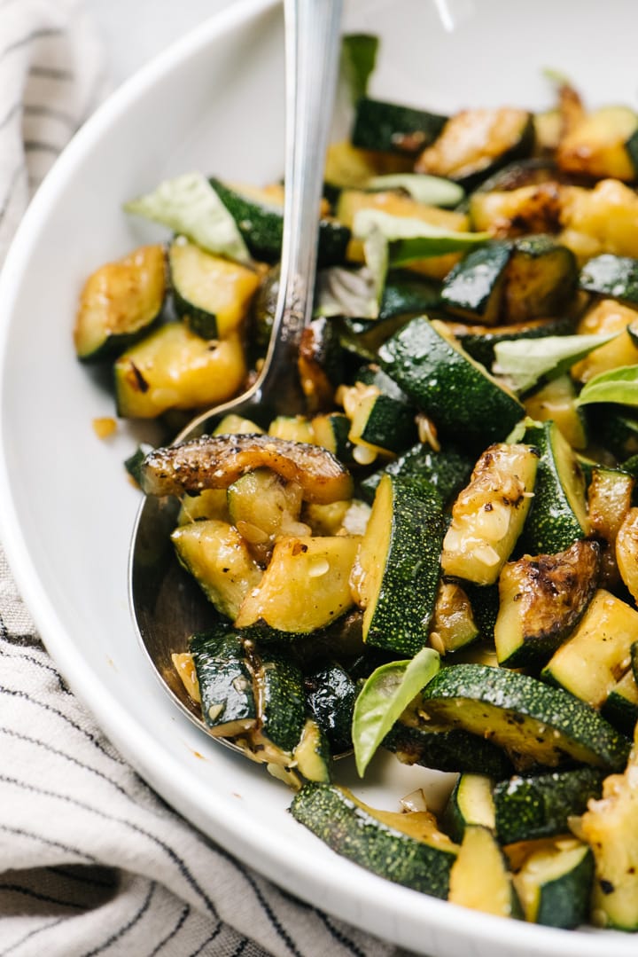 Side view, a silver serving spoon tucked into a white bowl of sautéed zucchini.